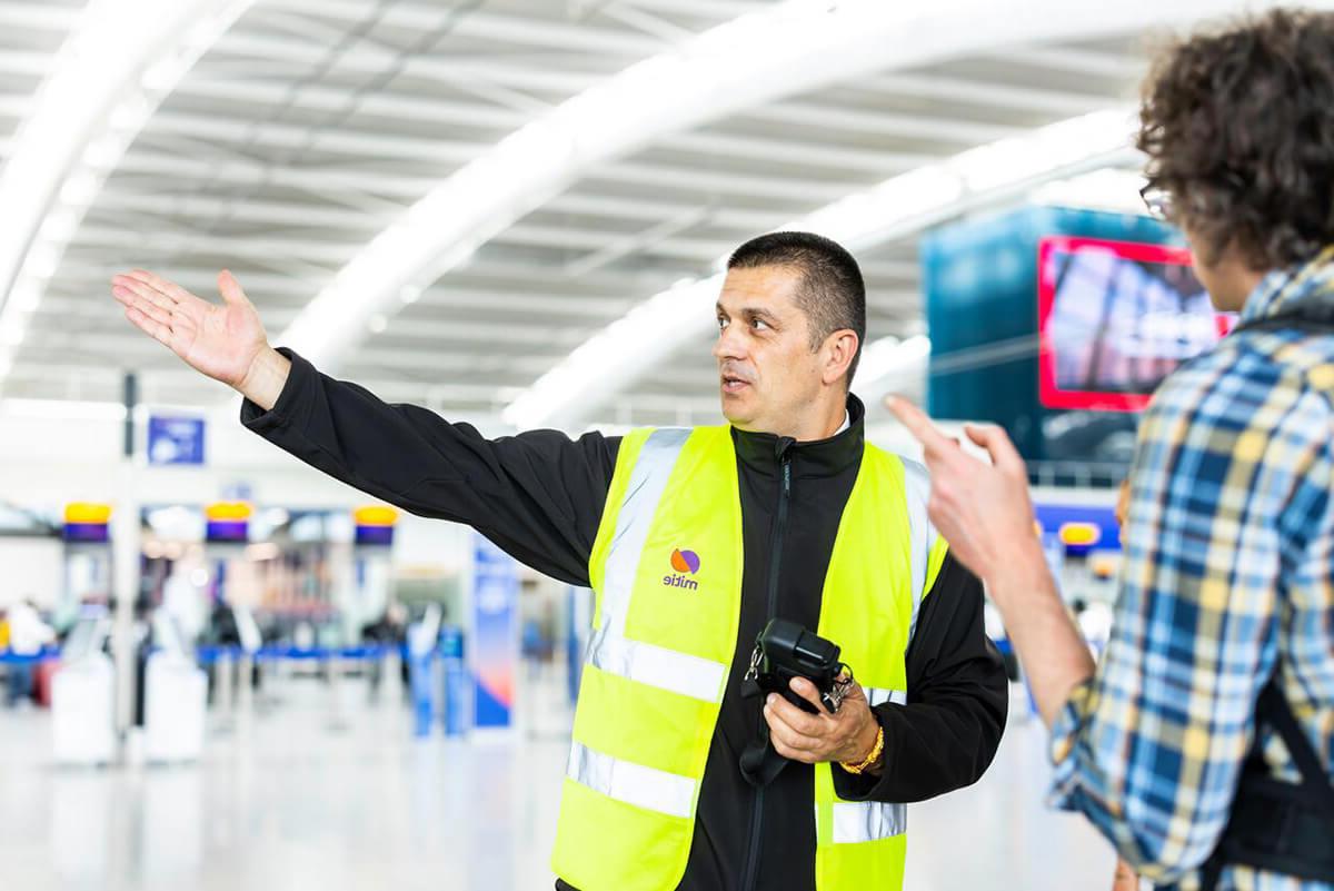 希思罗机场的男性保安, 戴着米蒂牌的高可见度, 指向右边，引导顾客