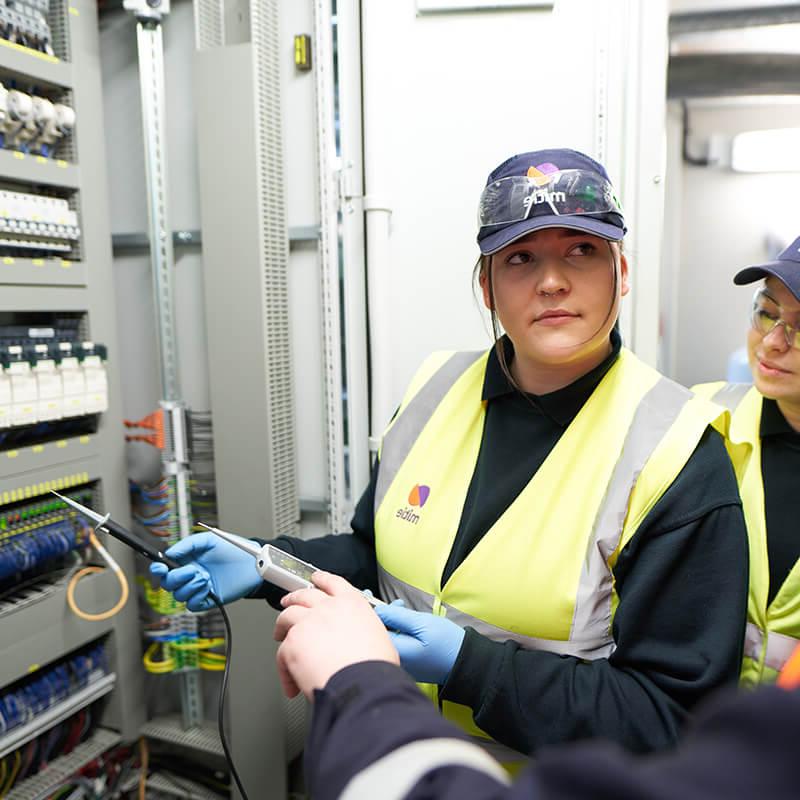 两个女工程师学徒, 穿着Mitie高能见度背心和帽子, 在一堵电线和保险丝墙前